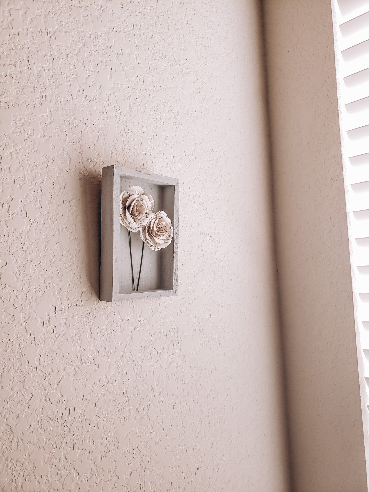 a pair of book page paper roses in a portrait shaped frame on a wall - Novel Blossoms Co