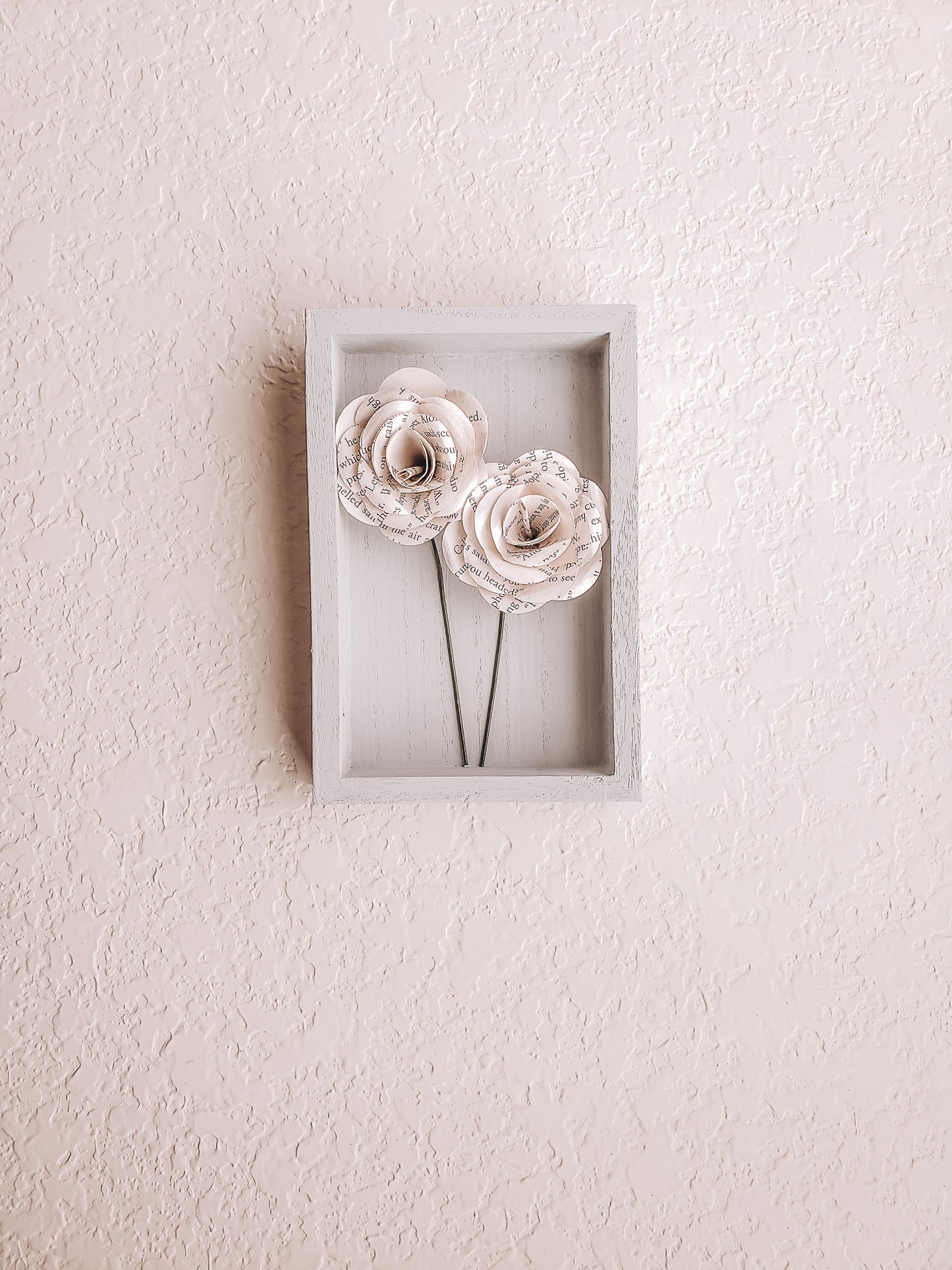 a pair of book page paper roses in a portrait shaped frame on a wall - Novel Blossoms Co