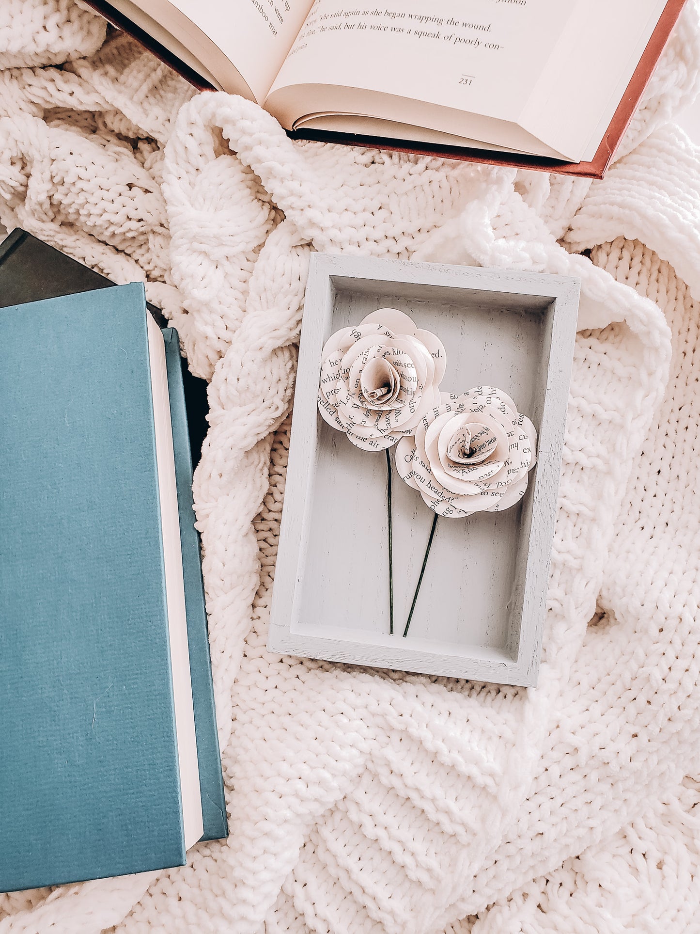 framed paper flowers made from book pages lying on a cable-knit sweater blanket and books scattered around - Novel Blossoms Co