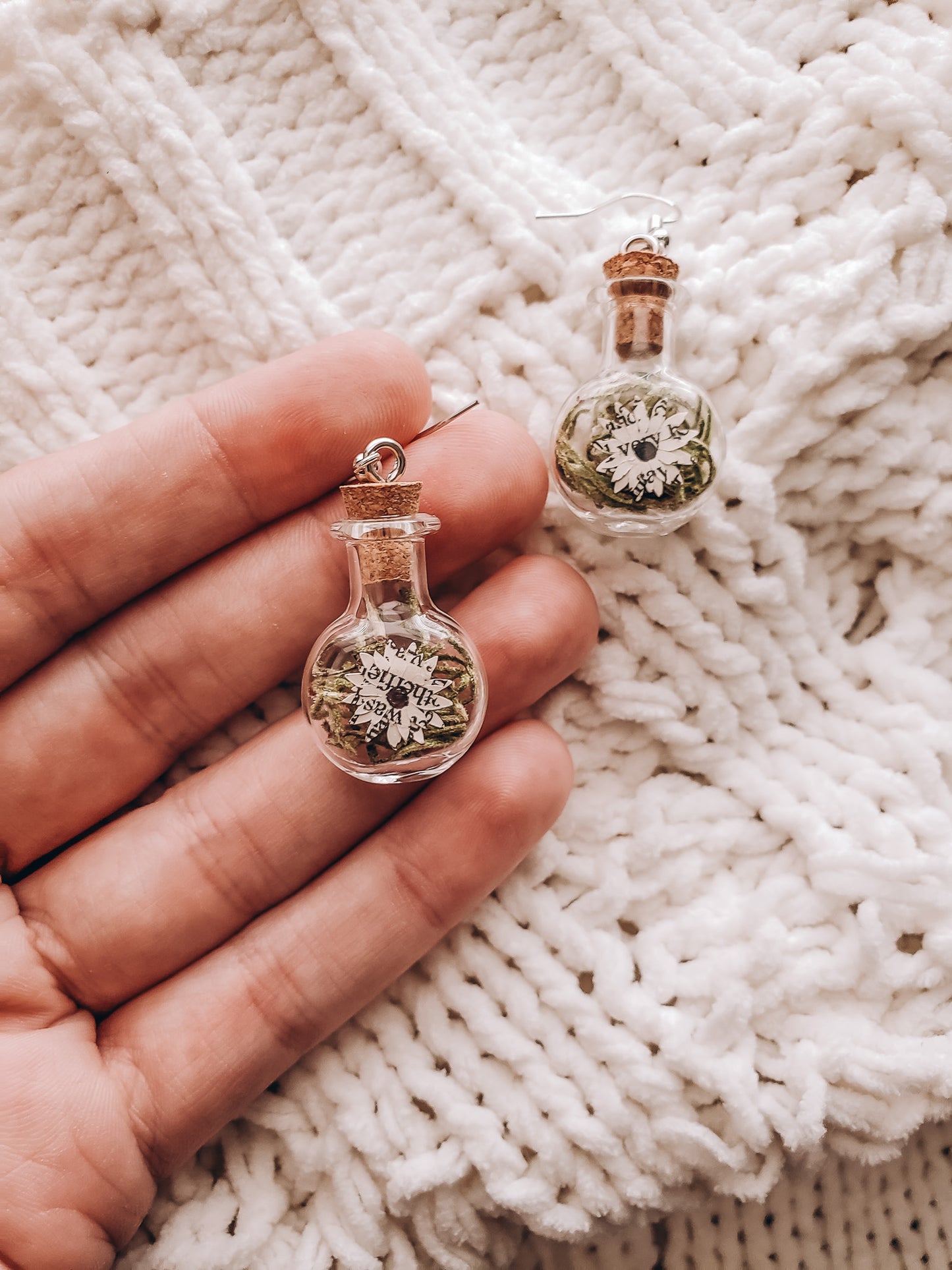 a hand holding a paper book page flower in tiny glass jar earrings on top of a cozy blanket - Novel Blossoms Co